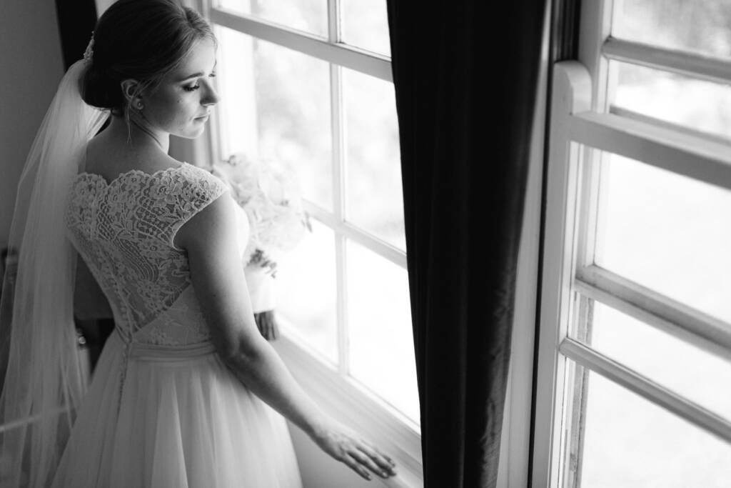 Veiled at a sash window between dark curtains, Sue rests a hand on the sill.