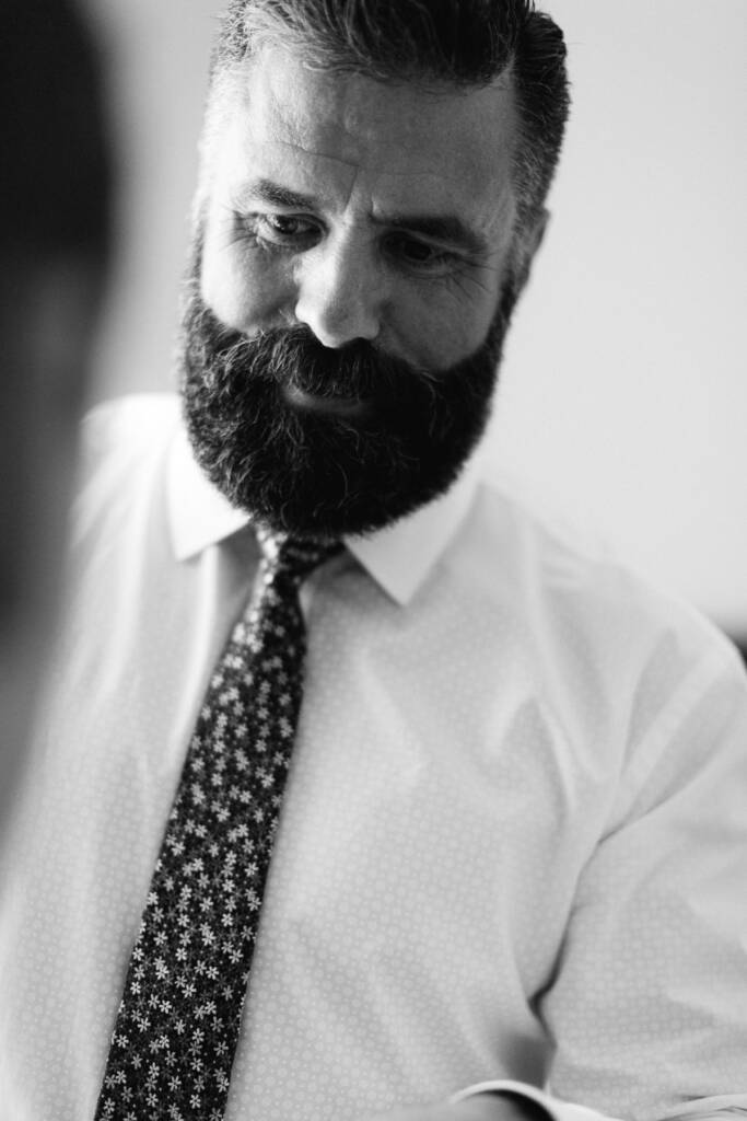 A bearded man in a white shirt and small floral tie looks down with a slight smile.