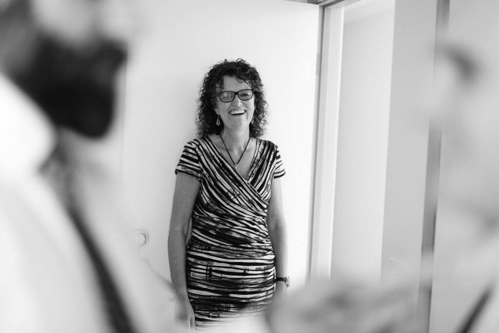A woman with curly hair and glasses laughs in a doorway, framed between two blurred men.