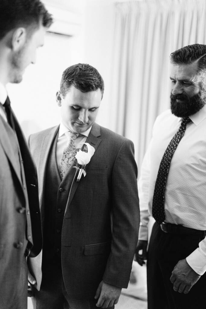 Chris looks down at the white rose boutonniere on his grey lapel, flanked by two men.
