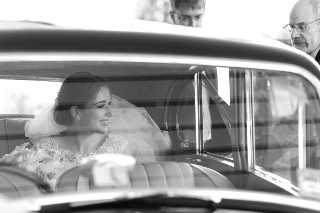 Veiled and smiling through the window of a vintage car, Sue holds her bouquet on her lap.