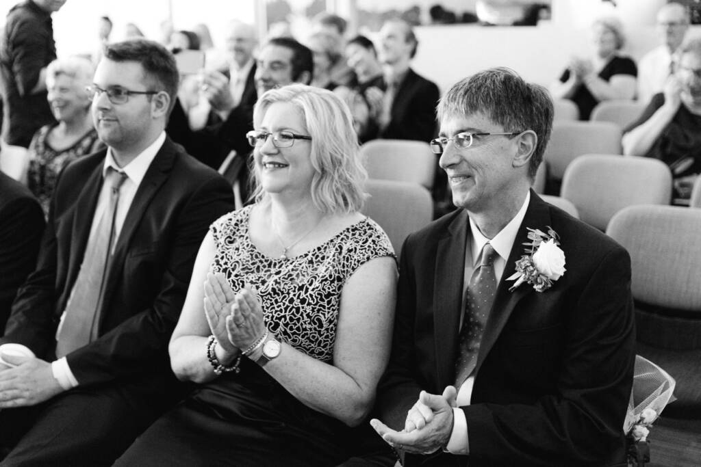 Front row guests applaud, a woman in glasses and a patterned top between two men in glasses.