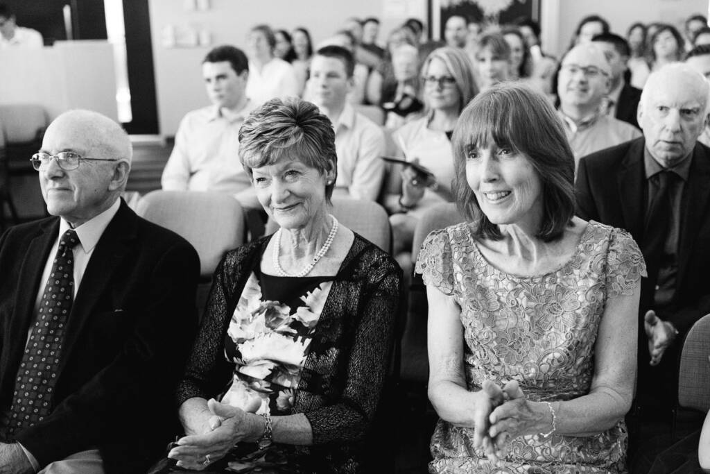 Three older guests in the front row smile and clap, one in pearls and a floral dress.