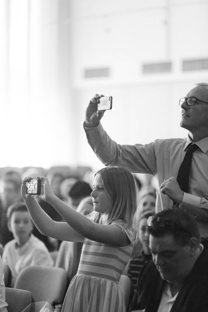 A girl in a striped dress and a man in glasses stand to film the ceremony on phones.