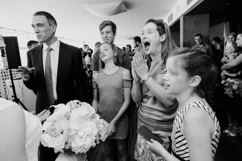 A woman in a satin top gasps with hands together as guests greet the couple after the ceremony.