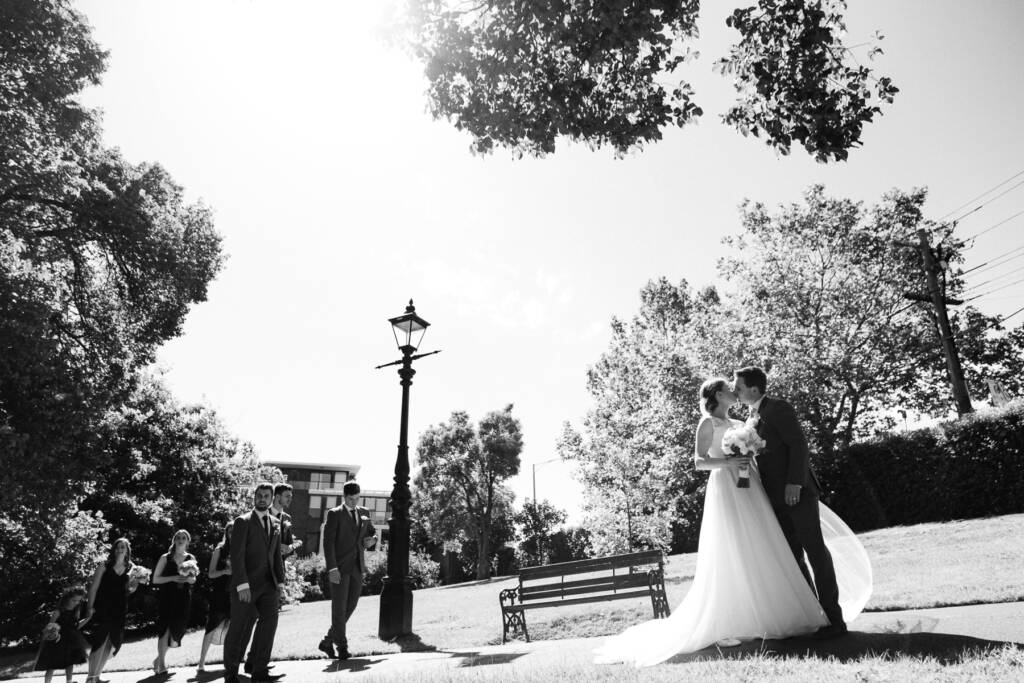 Sue and Chris kiss beside a Victorian lamp post in Canterbury Gardens as the bridal party walks in.