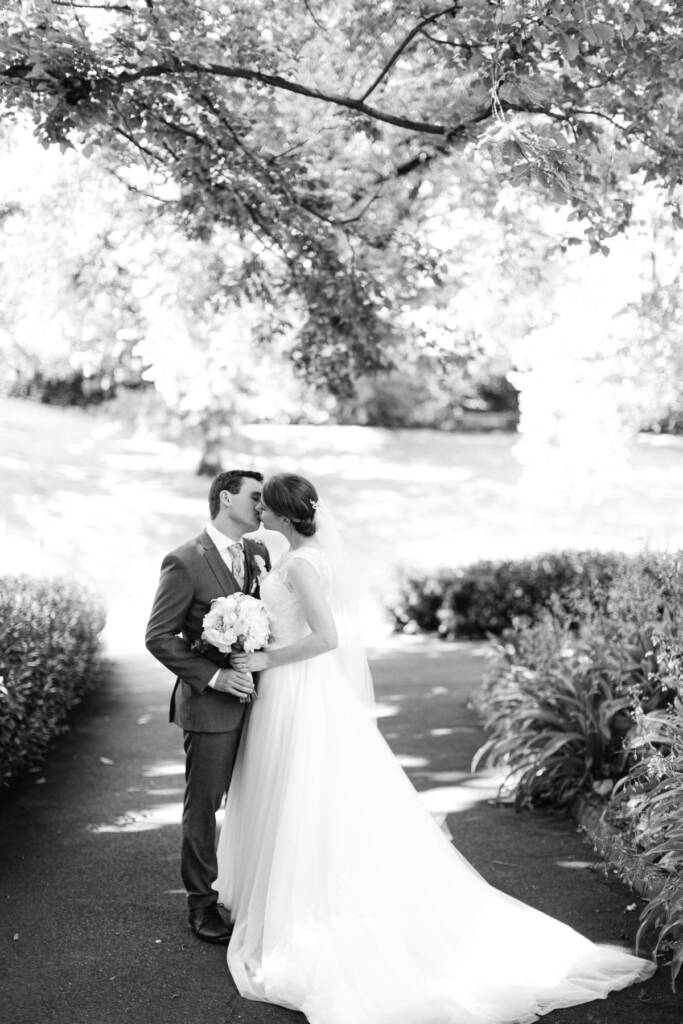 Sue and Chris kiss on a tree-lined path at Canterbury Gardens, her train spread behind them.