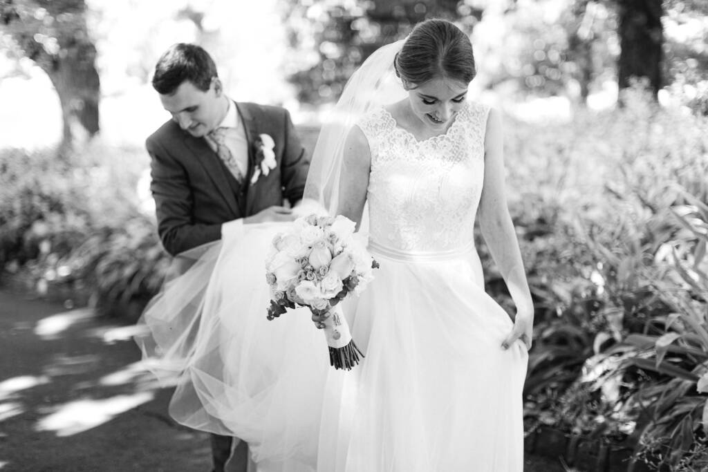 Chris carries Sue's veil and train as she walks with her bouquet at Canterbury Gardens.