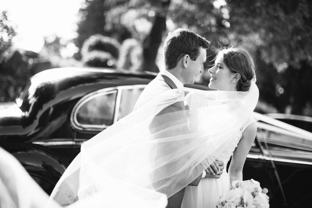 Sue's veil blows across Chris beside the classic car at Riversdale Golf Club's main entrance.
