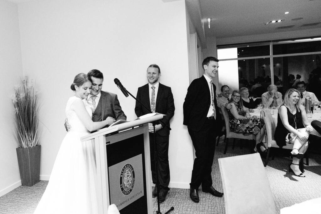 Sue and Chris look down at notes at the Riversdale Golf Club lectern while guests laugh.