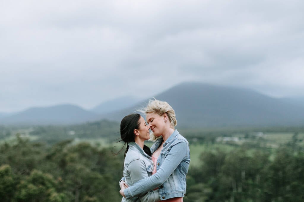 Ashlee and Kate embrace during their engagement shoot at Riverstone Estate, with cloud-covered hills behind them.