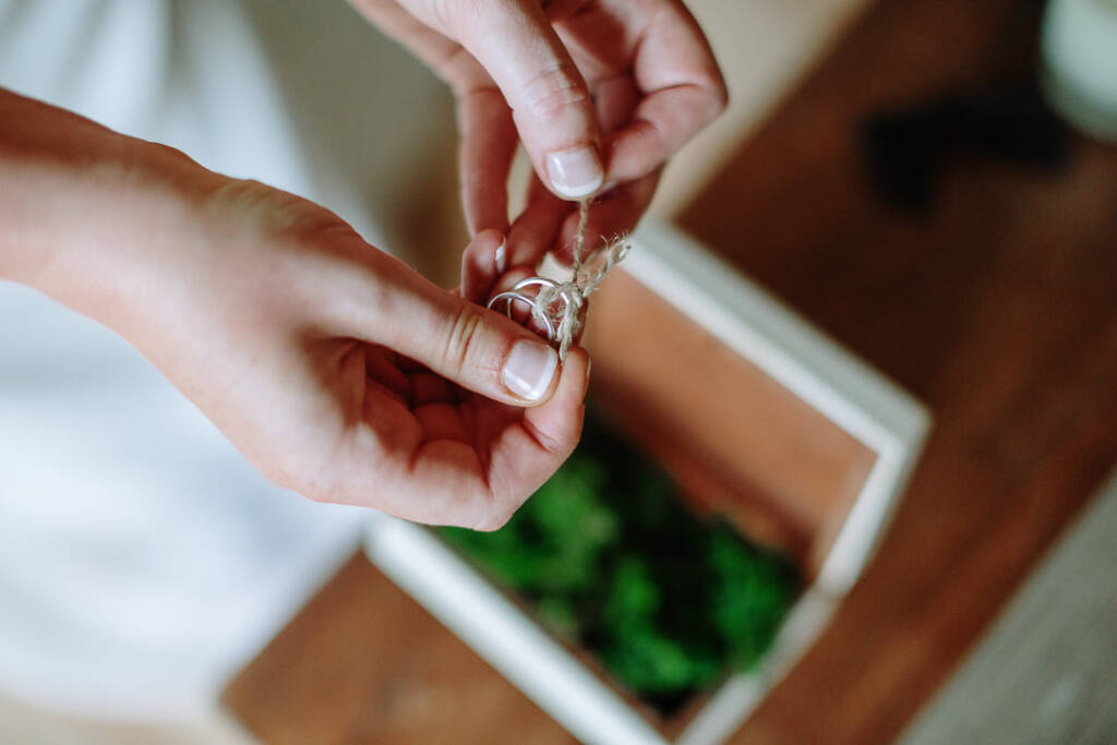 Hands hold two rings tied with twine above a moss-lined wooden ring box.