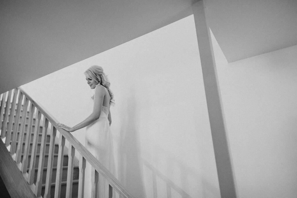 Kate stands on a timber staircase at her Chum Creek Airbnb in a wide black-and-white bridal portrait.