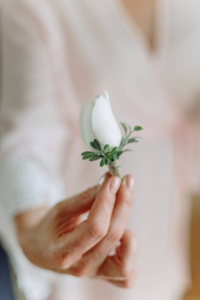 Hands hold a white rosebud buttonhole with a twine-wrapped stem.