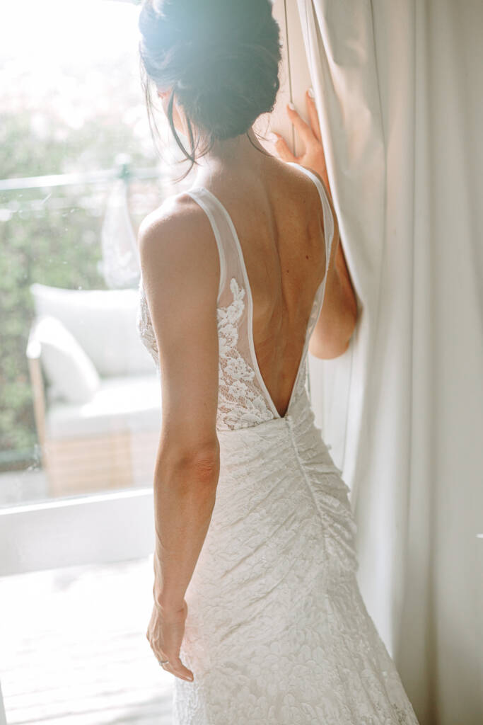Ashlee stands at her Healesville Airbnb's glass door, showing the low back of her lace gown.