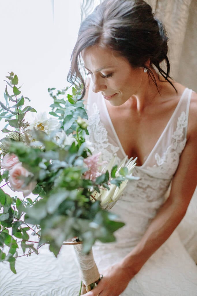 Ashlee looks down at her bouquet while seated beside the window at Riverstone Estate.