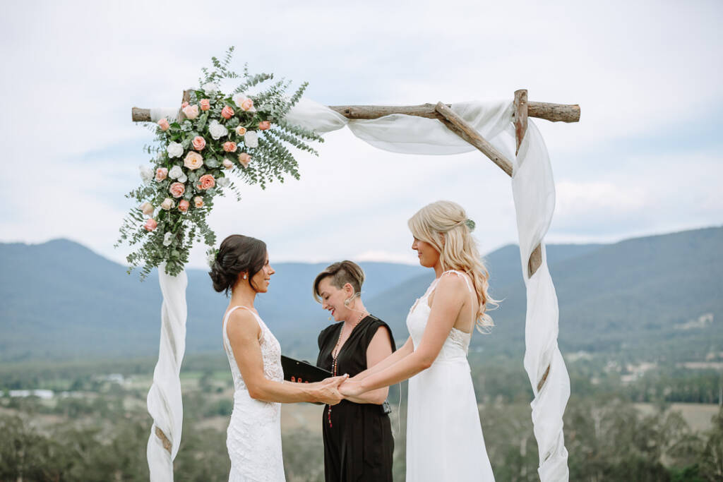 The celebrant reads from a folder between Ashlee and Kate beneath the arbour at Riverstone Estate.