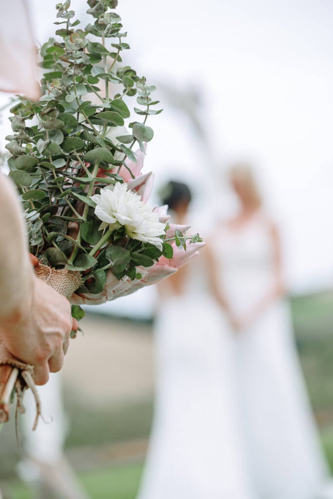 A bouquet fills the foreground with Ashlee and Kate beneath the arbour behind at Riverstone Estate.