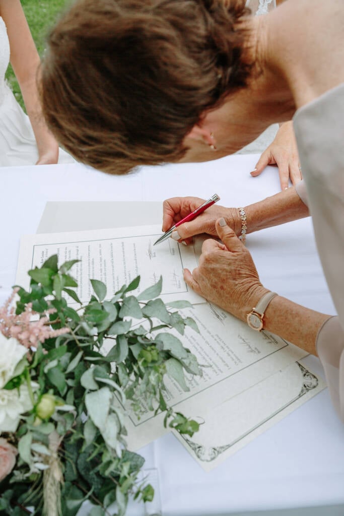 A witness signs the couple's commemorative ceremony document beside their bouquet at Riverstone.