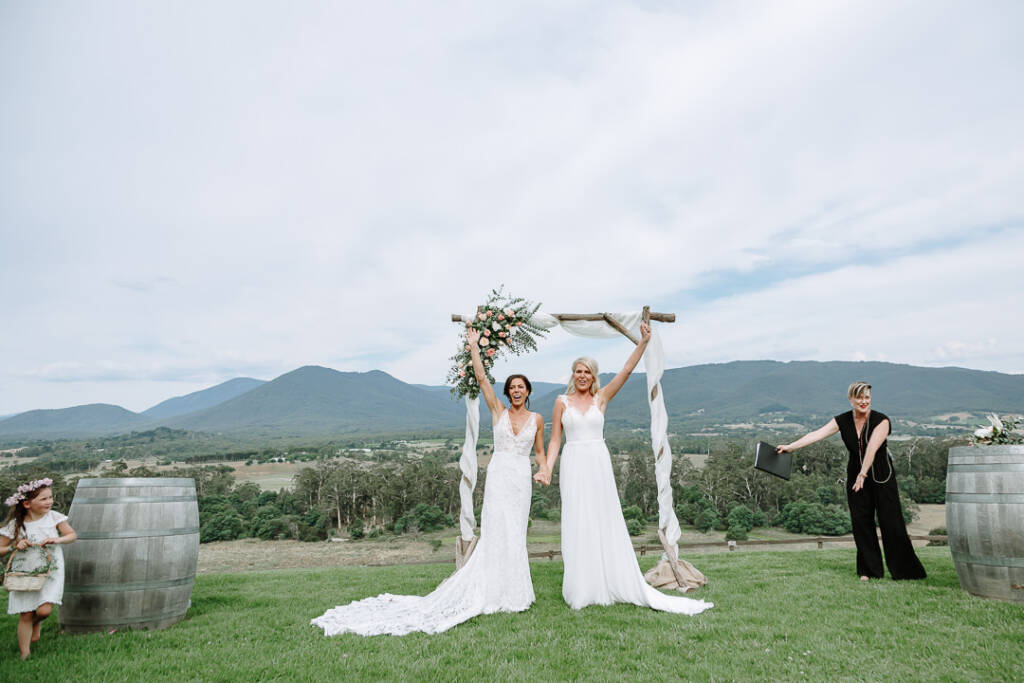 Ashlee and Kate raise their arms beneath the arbour at Riverstone Estate after their commitment ceremony.