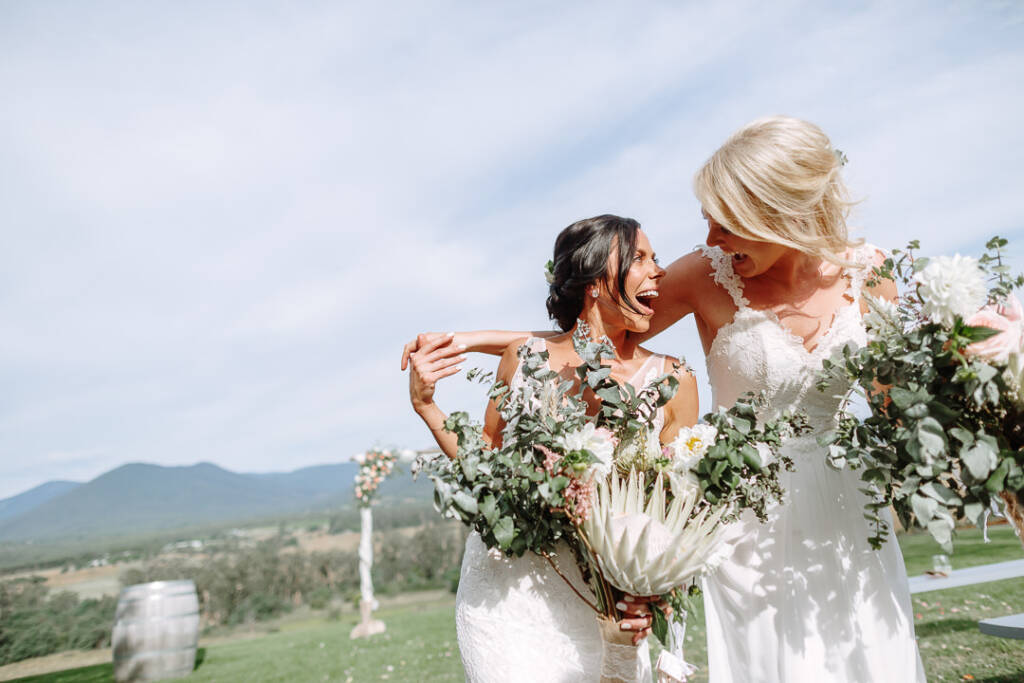 Ashlee and Kate laugh with their arms around each other, carrying bouquets at Riverstone Estate.