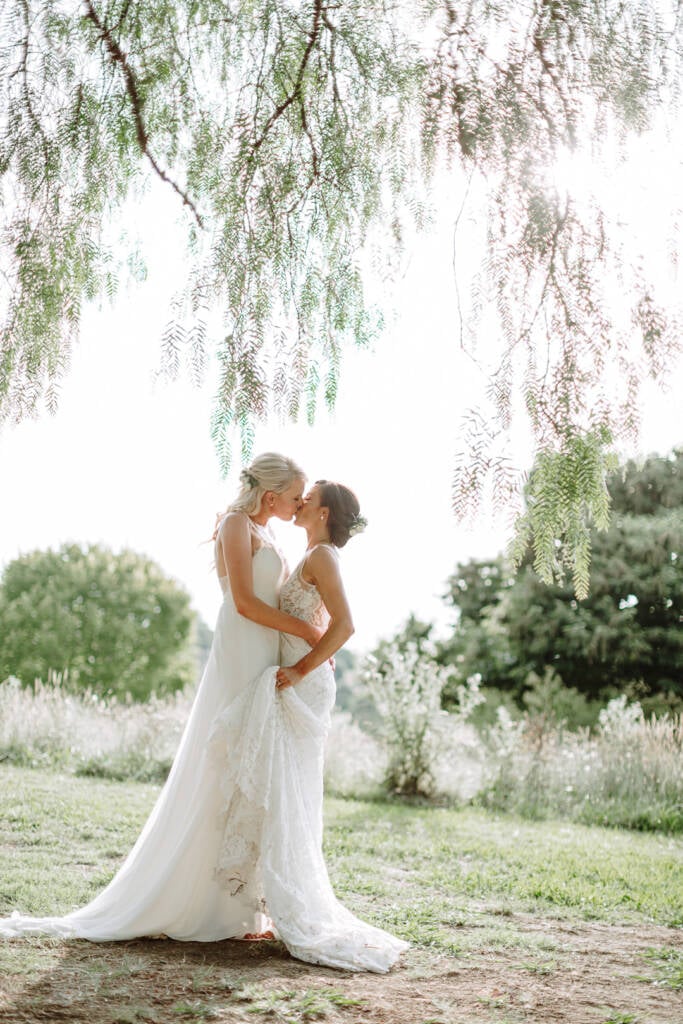 Ashlee and Kate kiss beneath sunlit branches at Riverstone.