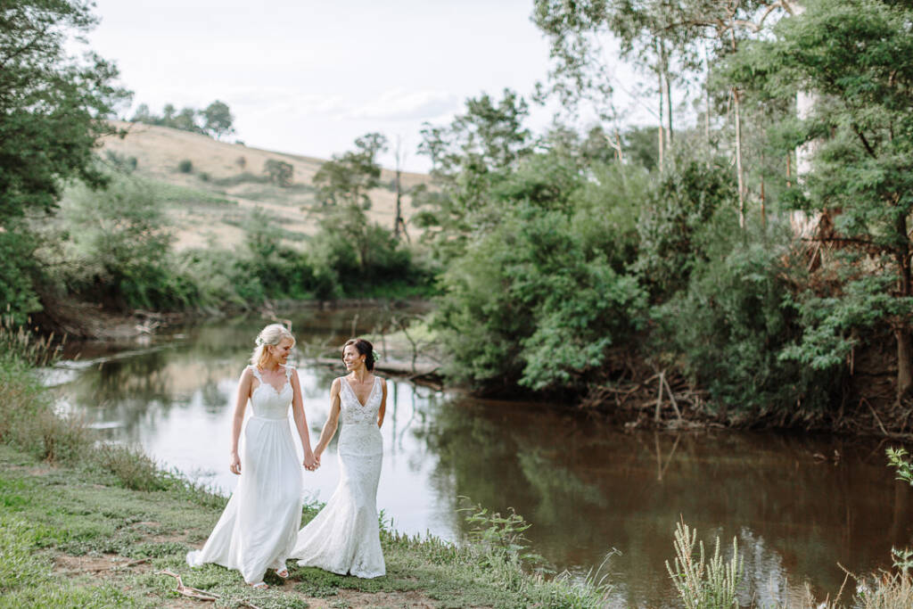 Ashlee and Kate hold hands beside the tree-lined river at Riverstone Estate in the Yarra Valley.