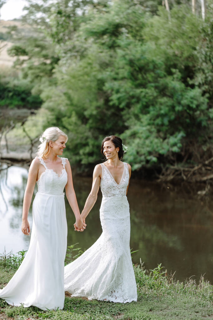 The couple smile at each other on the grassy riverbank at Riverstone.