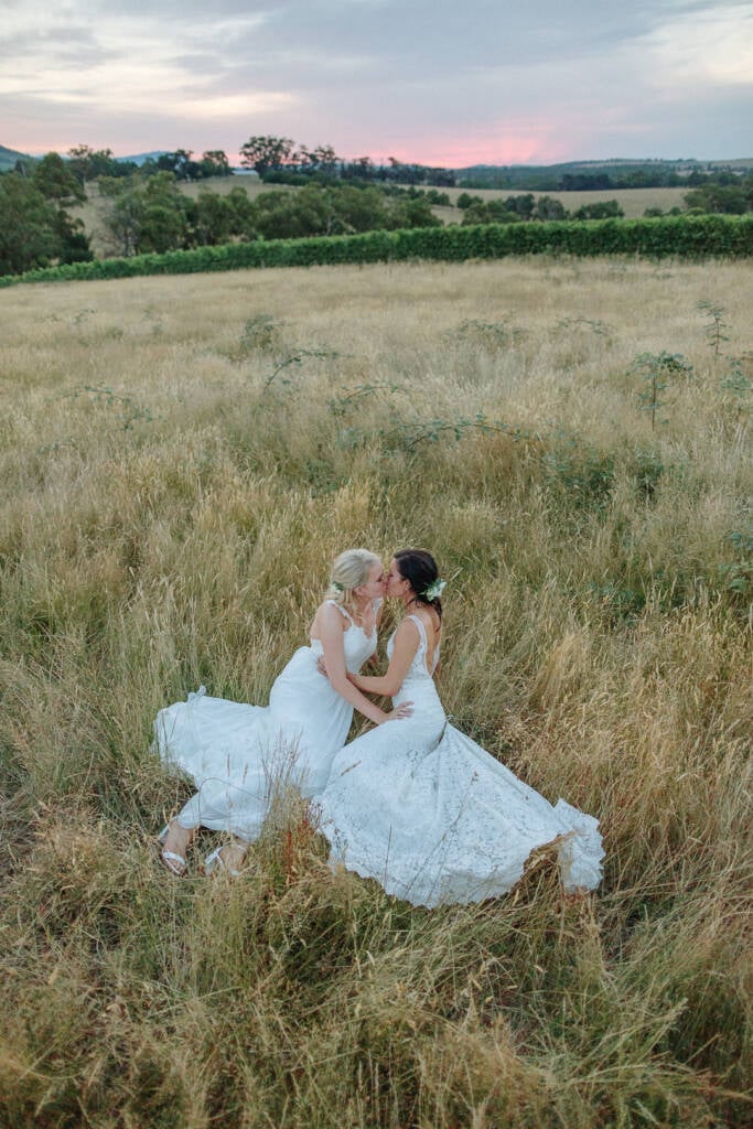 Ashlee and Kate kiss while seated in the grass beneath a pink sunset at Riverstone.