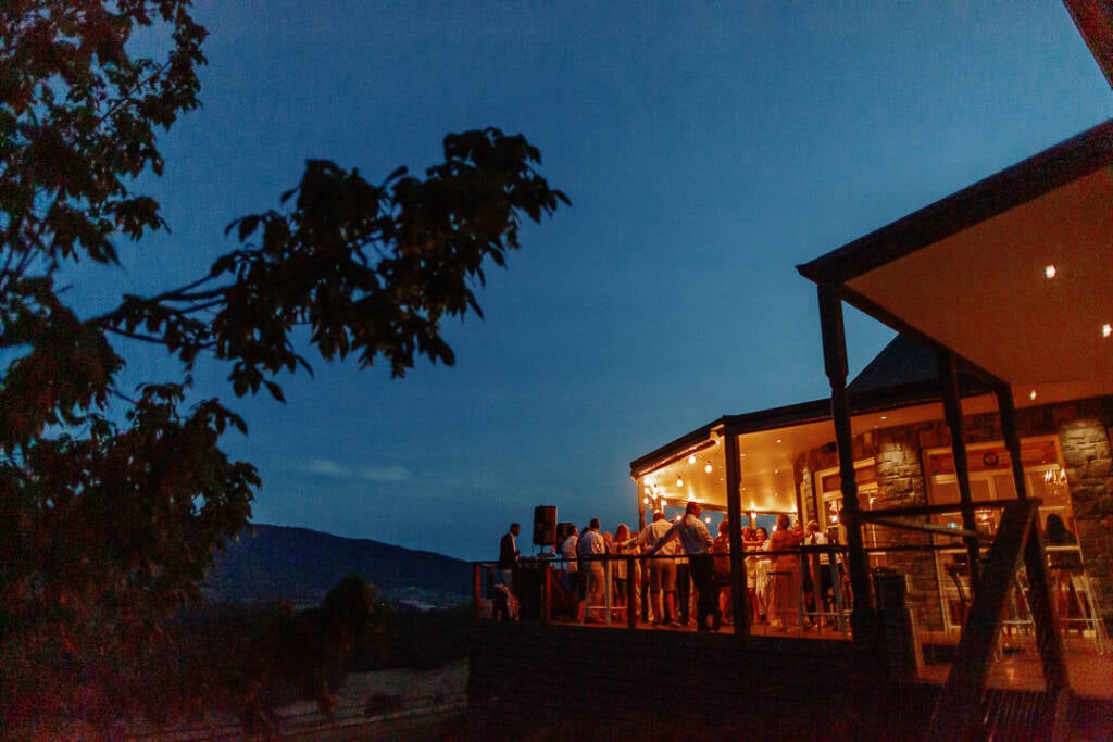 Riverstone's bluestone homestead and guest-filled verandah glow against the dark Yarra Valley hills.