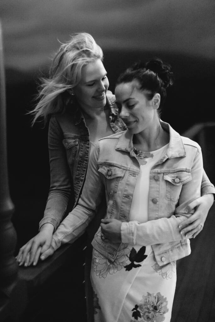 Black-and-white engagement portrait of Kate embracing Ashlee from behind in their casual jackets.