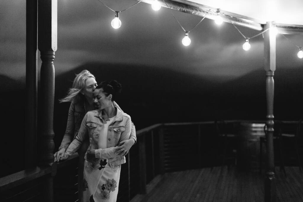 Kate kisses Ashlee's forehead beneath Riverstone Estate's verandah lights during their engagement shoot.