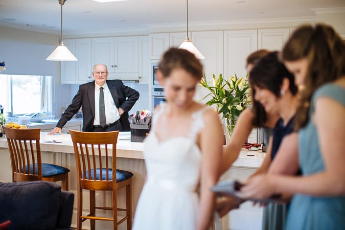 Frances waits as bridesmaids fasten her gown while a guest watches from behind the kitchen bench.