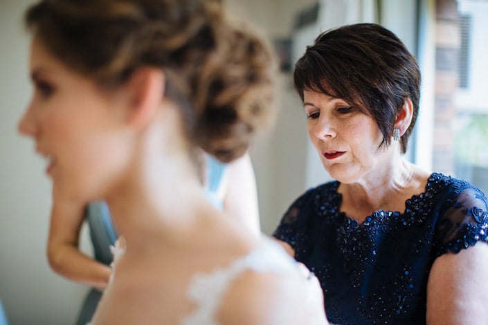 Close view of a woman in navy lace concentrating on the final fastening of Frances's gown.