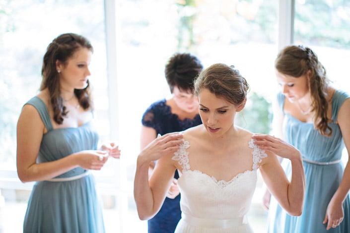 Two bridesmaids in dusty blue flank Frances as she settles the lace straps on her shoulders.