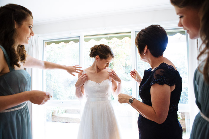 Bridesmaids adjust the straps of the gown in front of a bright window during the morning preparations.