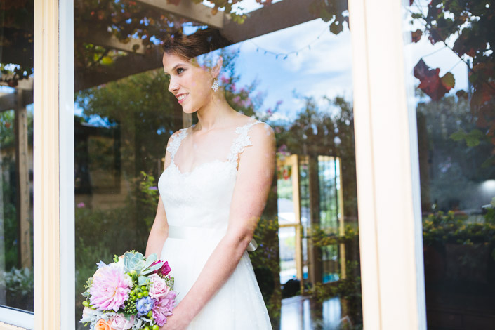 Frances smiles through a glazed door that reflects sky, trees and festoon lights, holding her bouquet.