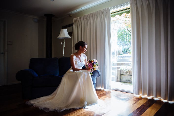 Seated on the arm of a navy sofa, Frances holds her pastel bouquet beside an open doorway.