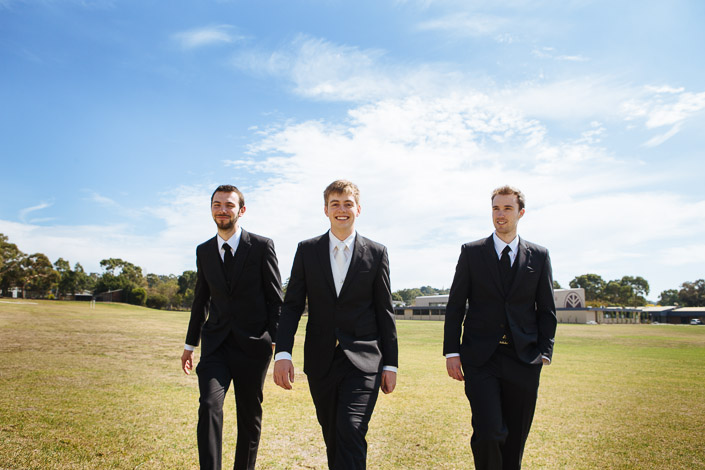 Tim and his groomsmen walk across a wide sunlit oval under a bright blue sky.