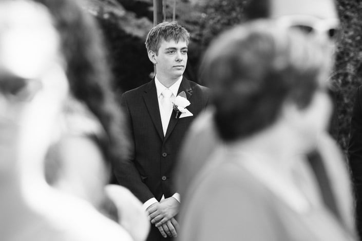 Black and white photo of Tim waiting at the ceremony, hands clasped, framed by guests.