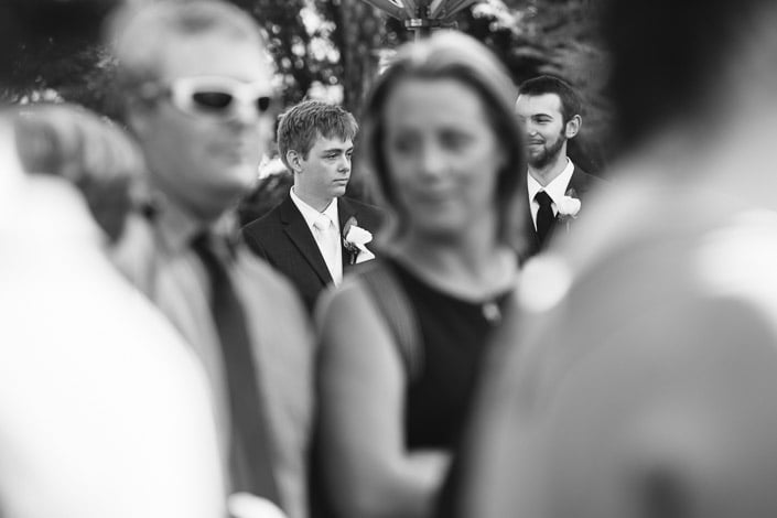 Black and white photo of a groomsman watching the ceremony through a blur of guests.