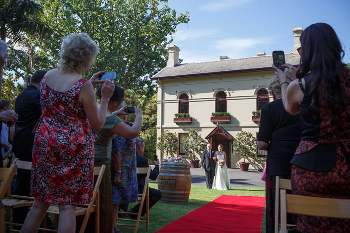 Guests raise phones as Frances walks the red carpet aisle at Gardens House, Royal Botanic Gardens.