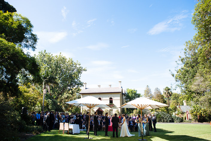 Wide view of the standing ceremony on the lawn at Gardens House with two market umbrellas.