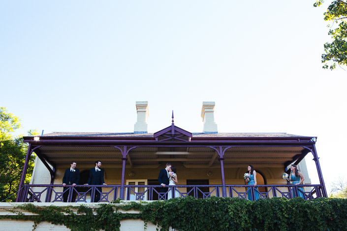 The wedding party lines a heritage verandah trimmed in plum-painted timber, spaced along the rail.