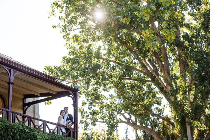Low sun flares through a huge tree as Frances and Tim stand together at a verandah rail.