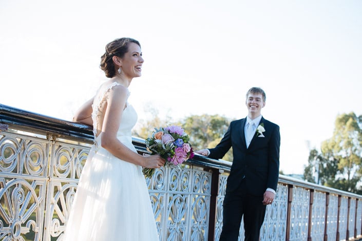 Frances laughs at the rail of Morell Bridge while Tim watches her from further along in golden light.