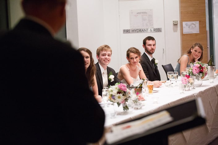 The bridal table listens to a speech, seen past the speaker's shoulder at the lectern.