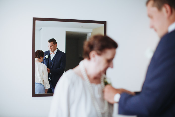 In the mirror, a woman in pearls helps thread the groom's white rose boutonniere.