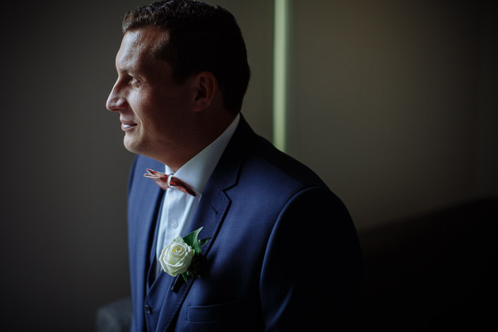 The groom's profile softens toward the light, blush bow tie crisp.