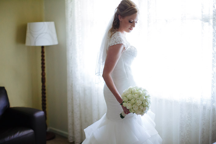 The bride looks down over the mermaid train by the lace curtains, roses in hand.